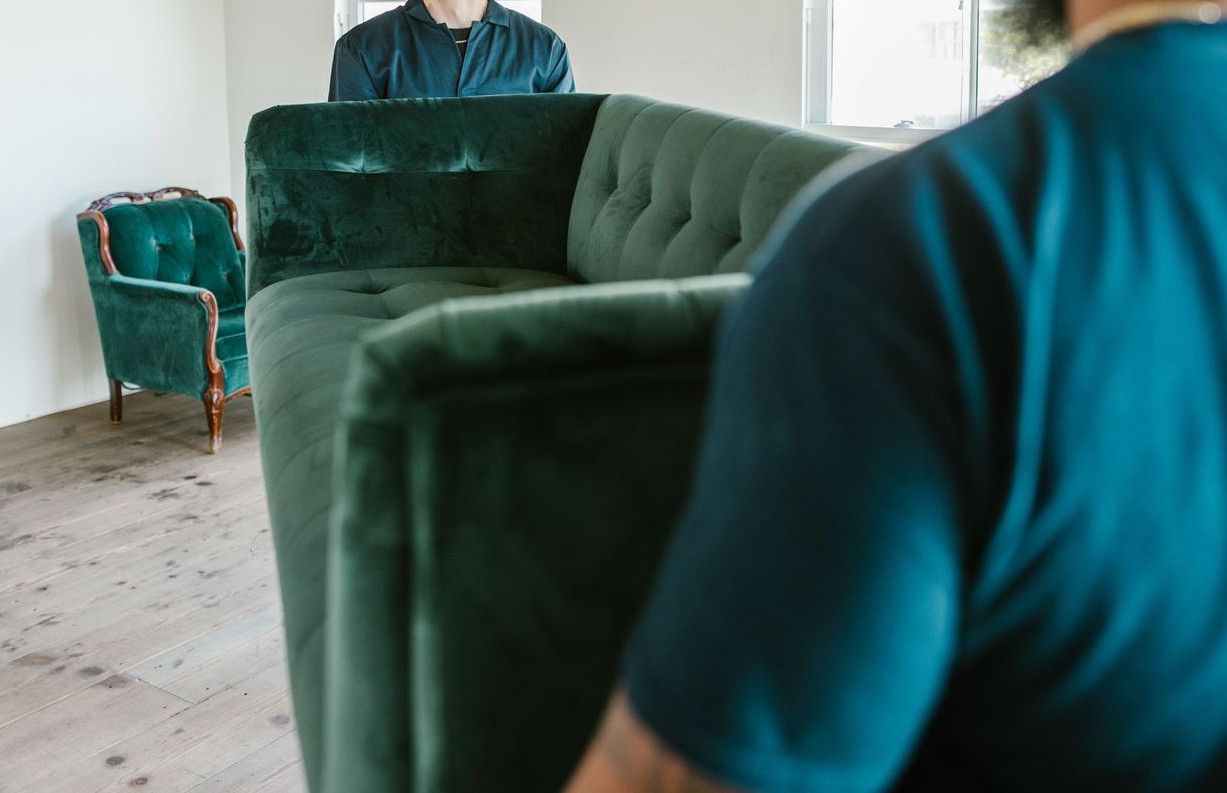 Two movers carrying a green couch in a sunlit room with packed boxes.